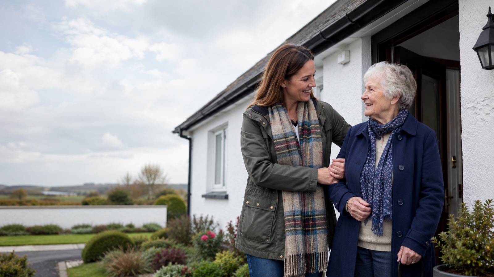 An adult daughter helping her older mother at the front door of a neat Irish bungalow